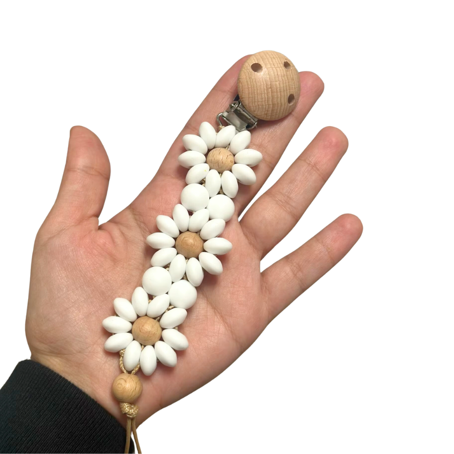 White Boho baby pacifier clip in the shape of a daisy flower held in a hand. Made of silicone saucer beads on a wooden clip with wooden beads for reborn dolls.