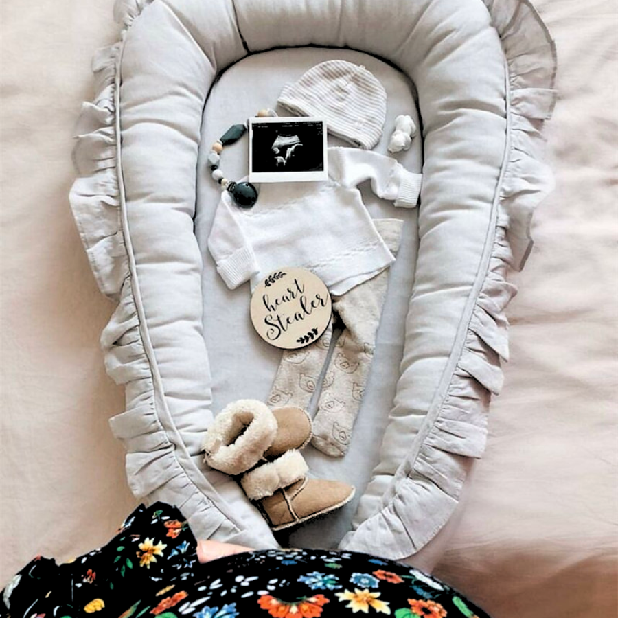 Pregnant woman holding her belly standing in front of a grey baby boy shooting star boho baby nest with ruffles for newborn babies and reborn doll displays.