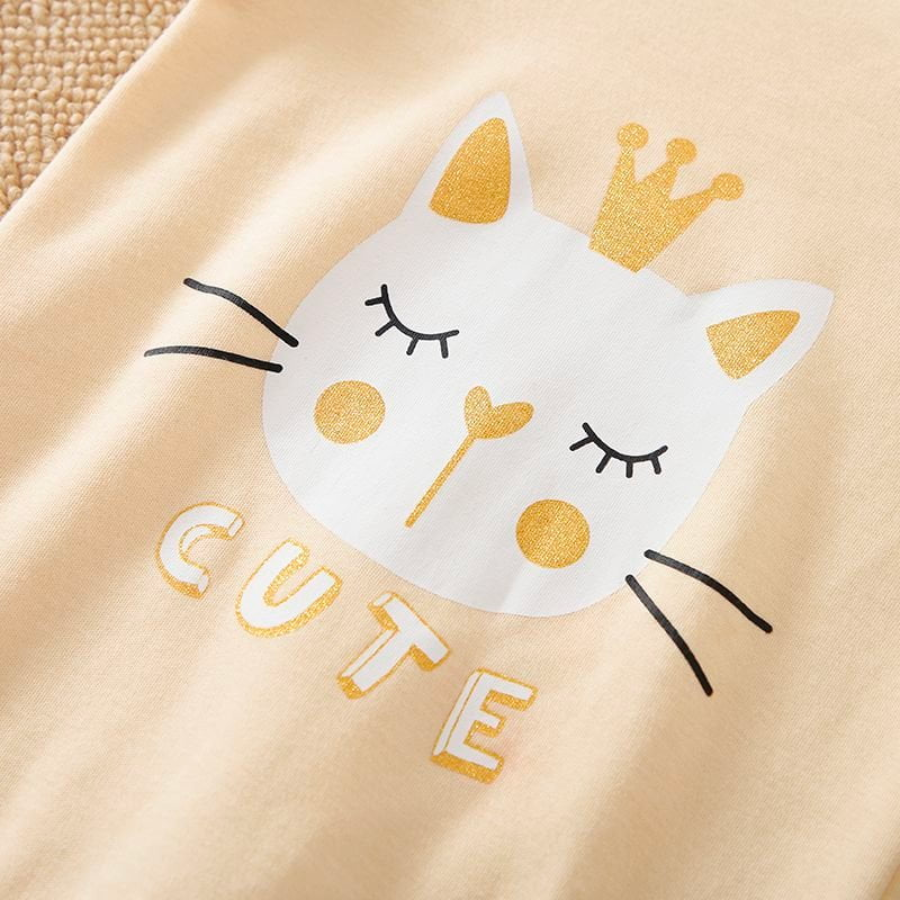Close-up of the cat face on a light yellow long-sleeve jumpsuit romper featuring a white cat with closed eyes and a gold crown on it's head and the word "cute" underneath it for reborn baby girl dolls.