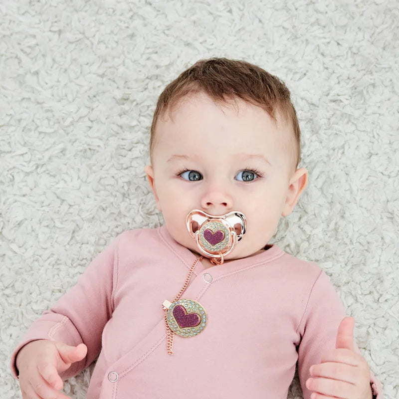 Newborn baby wearing a metallic pink Bling luxury rhinestone pacifiers and matching gold chain clips.