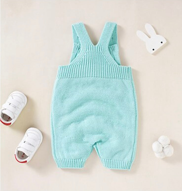 Back of aqua blue knitted overall shorts for reborn dolls with a white silhouette of a bunny rabbit sticking out of the pocket paired with a white long-sleeve onesie.
