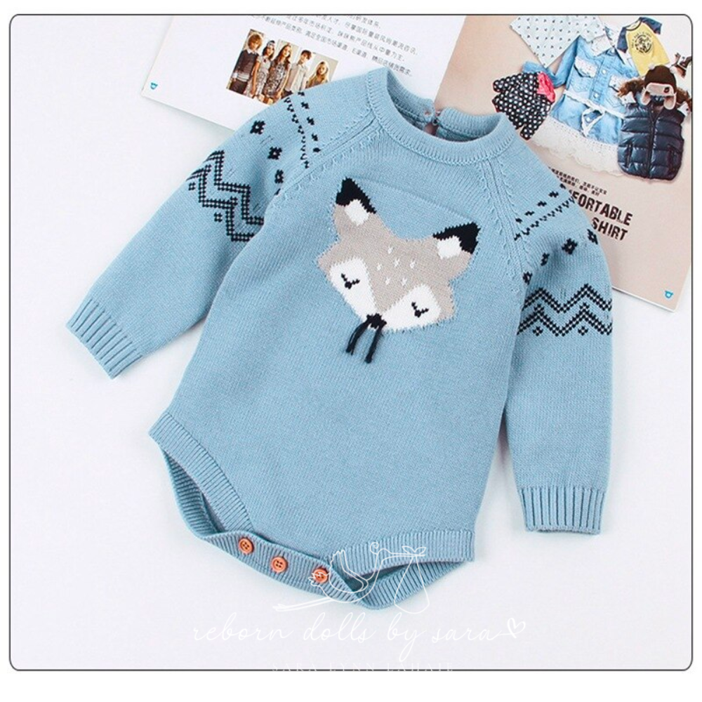 Blue knitted long-sleeve sweater onesie for reborn baby dolls with fox face knitted on the front who has black whiskers made of string. The sleeves feature black chevrons and diamond detail. The back of the romper has a button on it as does the crotch of the romper.