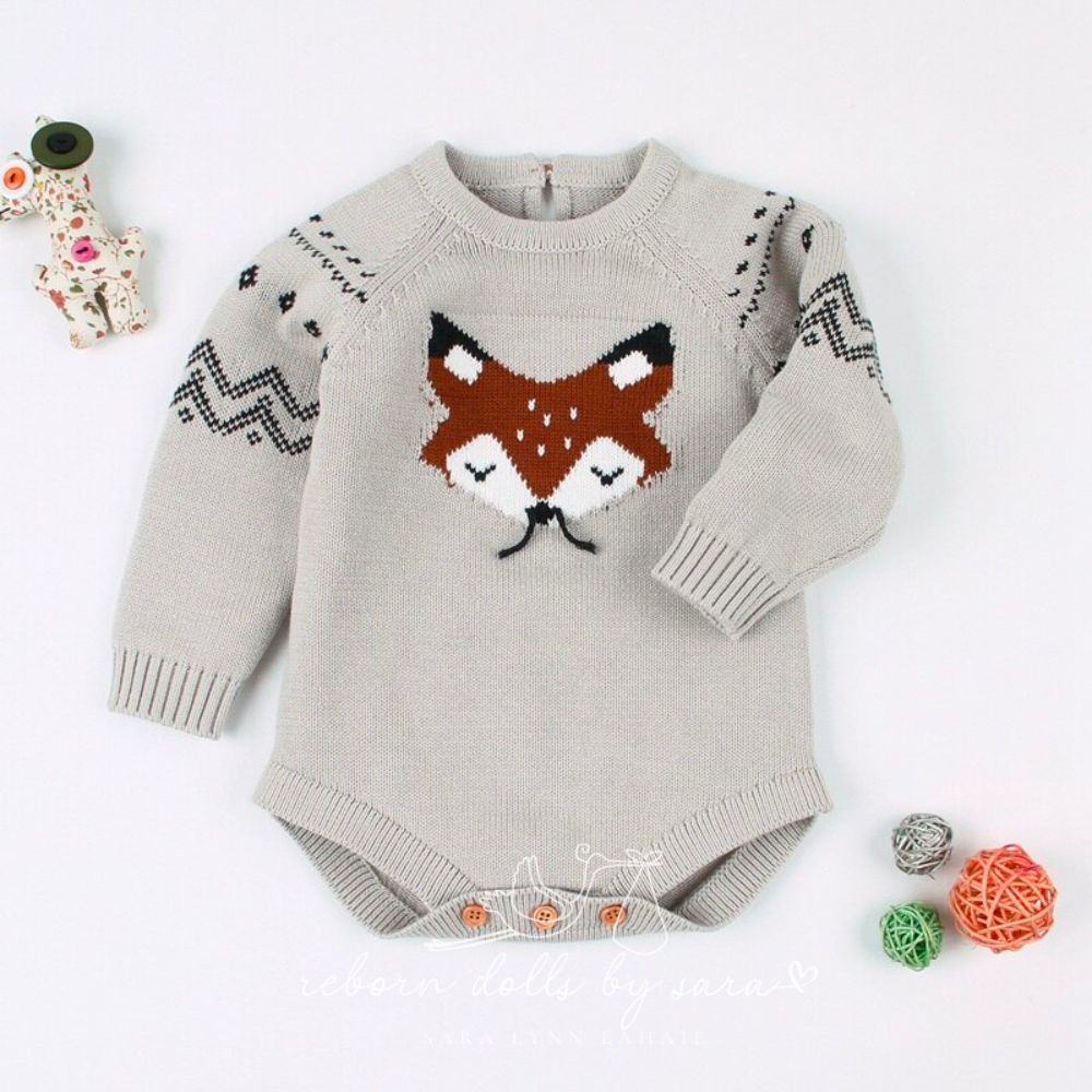 Grey knitted long-sleeve sweater onesie for reborn baby dolls with fox face knitted on the front who has black whiskers made of string. The sleeves feature black chevrons and diamond detail. The back of the romper has a button on it as does the crotch of the romper.