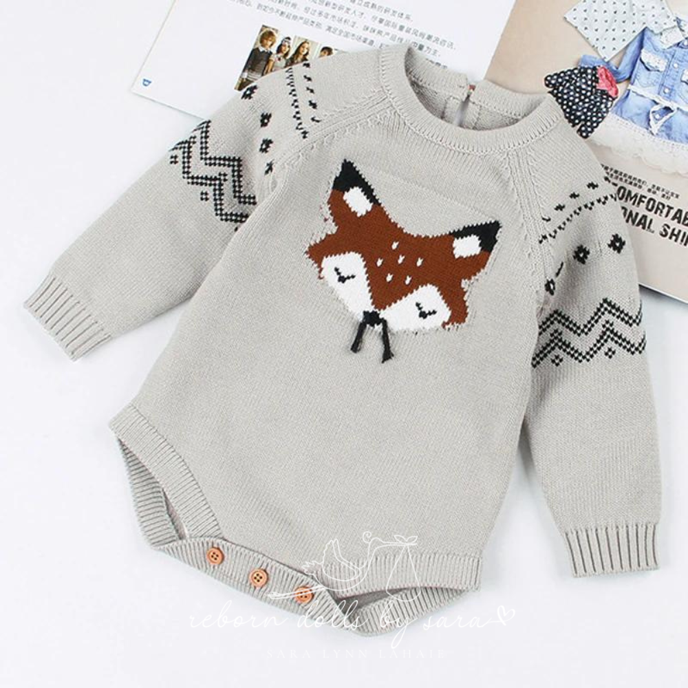 Grey knitted long-sleeve sweater onesie for reborn baby dolls with fox face knitted on the front who has black whiskers made of string.  The sleeves feature black chevrons and diamond detail.  The back of the romper has a button on it as does the crotch of the romper.  