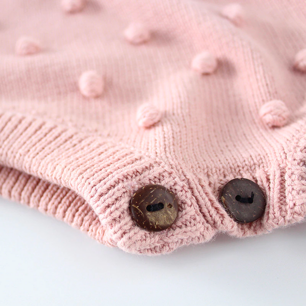 Close-up of crotch buttons on the Baby pink sleeveless knitted bubble romper with pink pompoms on the front, buttons on the shoulders, and buttons at the crotch. It also features a functional drawstring and is made to fit newborns baby girls to 2 year olds and reborn dolls.