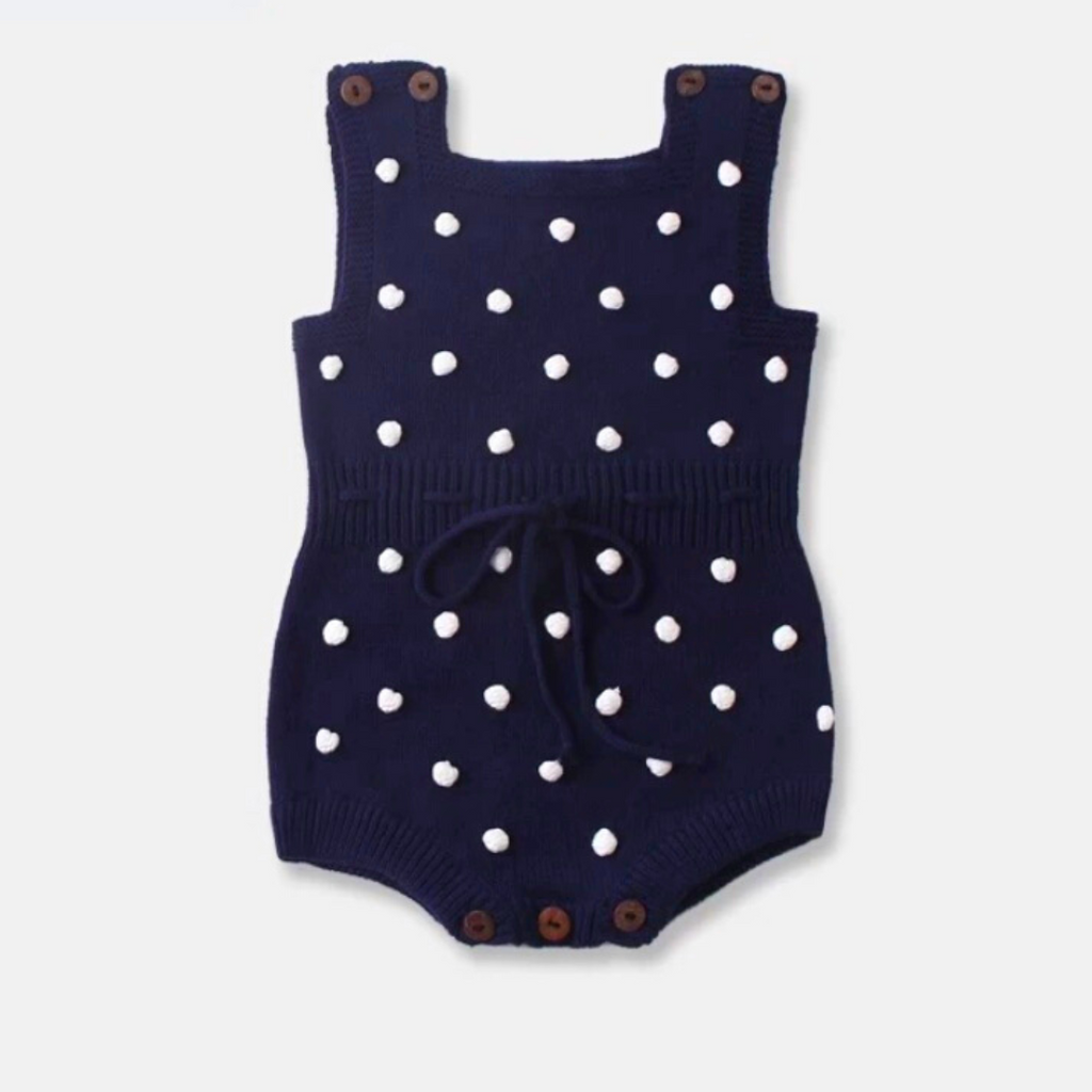 navy blue sleeveless knitted bubble romper with white pompoms on the front, buttons on the shoulders, and buttons at the crotch. It also features a functional drawstring and is made to fit newborns baby girls to 2 year olds and reborn dolls.