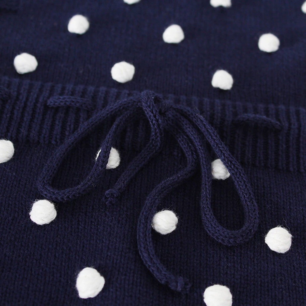 Close-up of the drawstring on the navy blue sleeveless knitted bubble romper with white pompoms on the front, buttons on the shoulders, and buttons at the crotch. It also features a functional drawstring and is made to fit newborns baby girls to 2 year olds and reborn dolls.