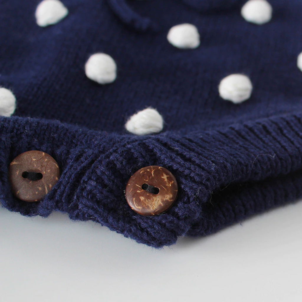 Close-up of crotch buttons on the navy blue sleeveless knitted bubble romper with white pompoms on the front, buttons on the shoulders, and buttons at the crotch. It also features a functional drawstring and is made to fit newborns baby girls to 2 year olds and reborn dolls.