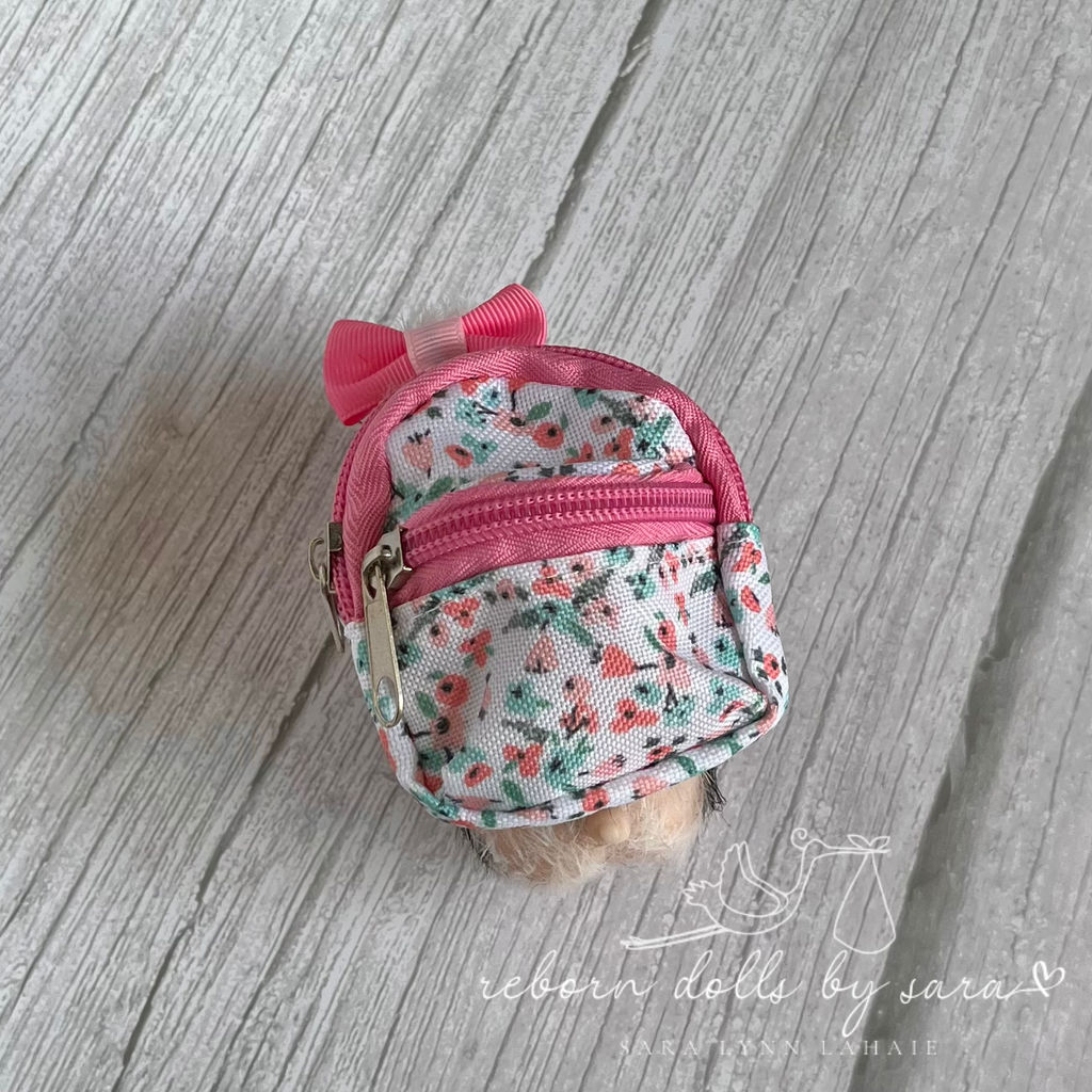 Miniature silicone piglet wearing a miniature pink and floral backpack.