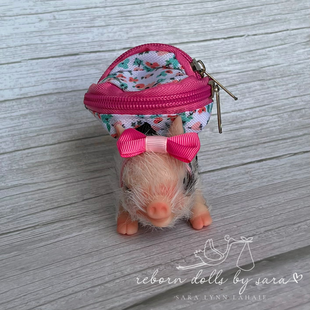 Black and white piglet wearing a hot pink and floral miniature backpack and a pink bow in it's hair.