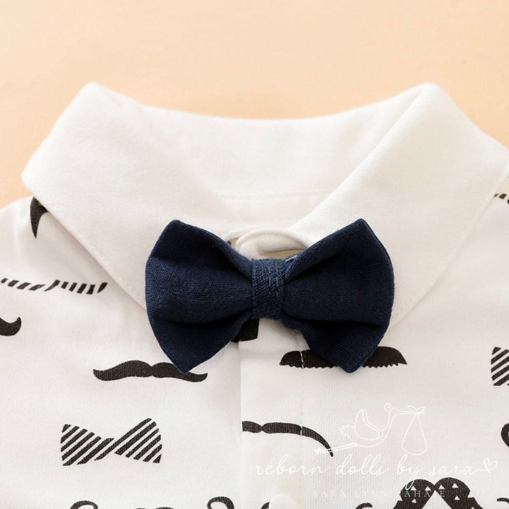 White long-sleeve button-up collared romper with black moustaches all over it and a navy blue bow tie for newborn babies and reborn dolls. 