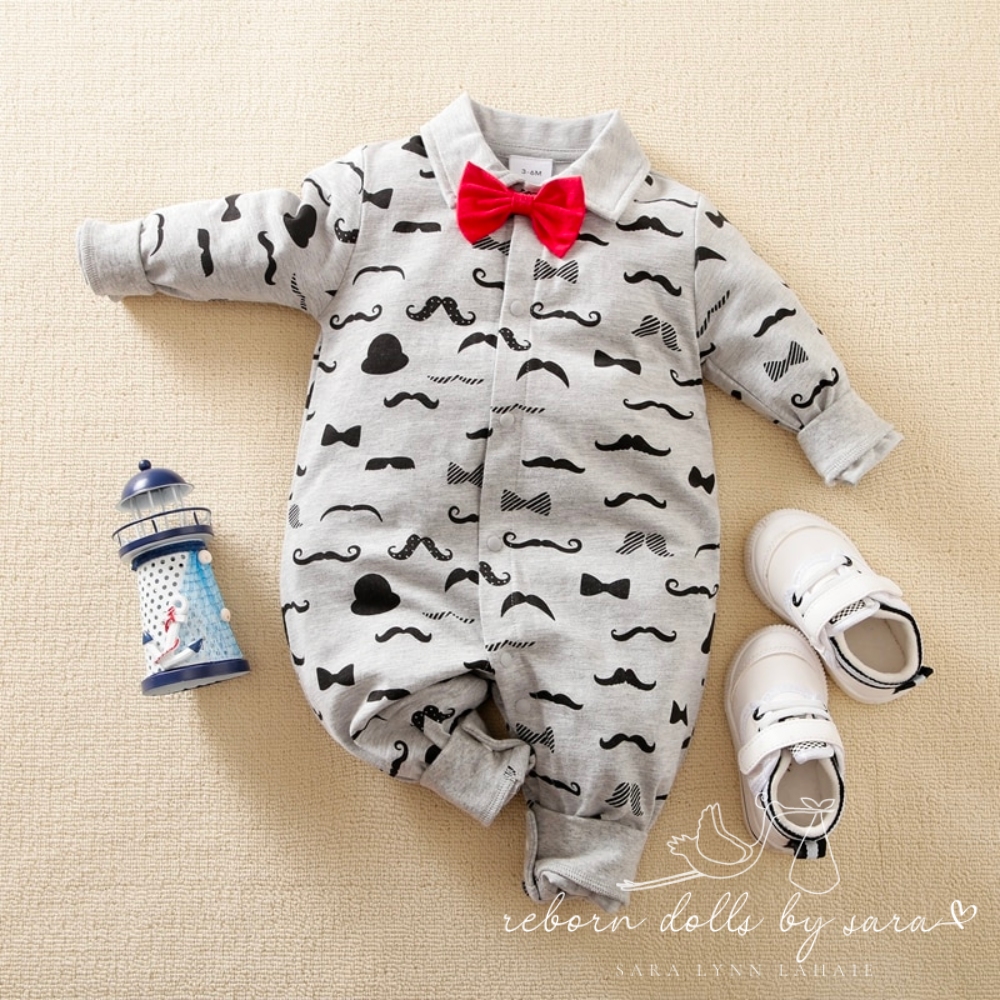 Grey long-sleeve button-up collared romper with black moustaches all over it and a red bow tie for newborn babies and reborn dolls. 