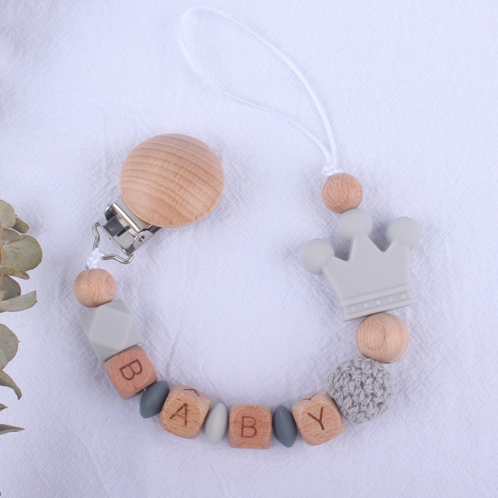 Light grey and dark grey personalized silicone pacifier clips with wooden fastener, crown bead, crochet beads, wooden beads with spacers between them, and silicone beads with baby's name for reborn baby dolls.