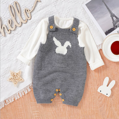 Grey knitted overall shorts for reborn dolls with a white silhouette of a bunny rabbit sticking out of the pocket. The overalls have been paired with a white long-sleeve onesie underneath.
