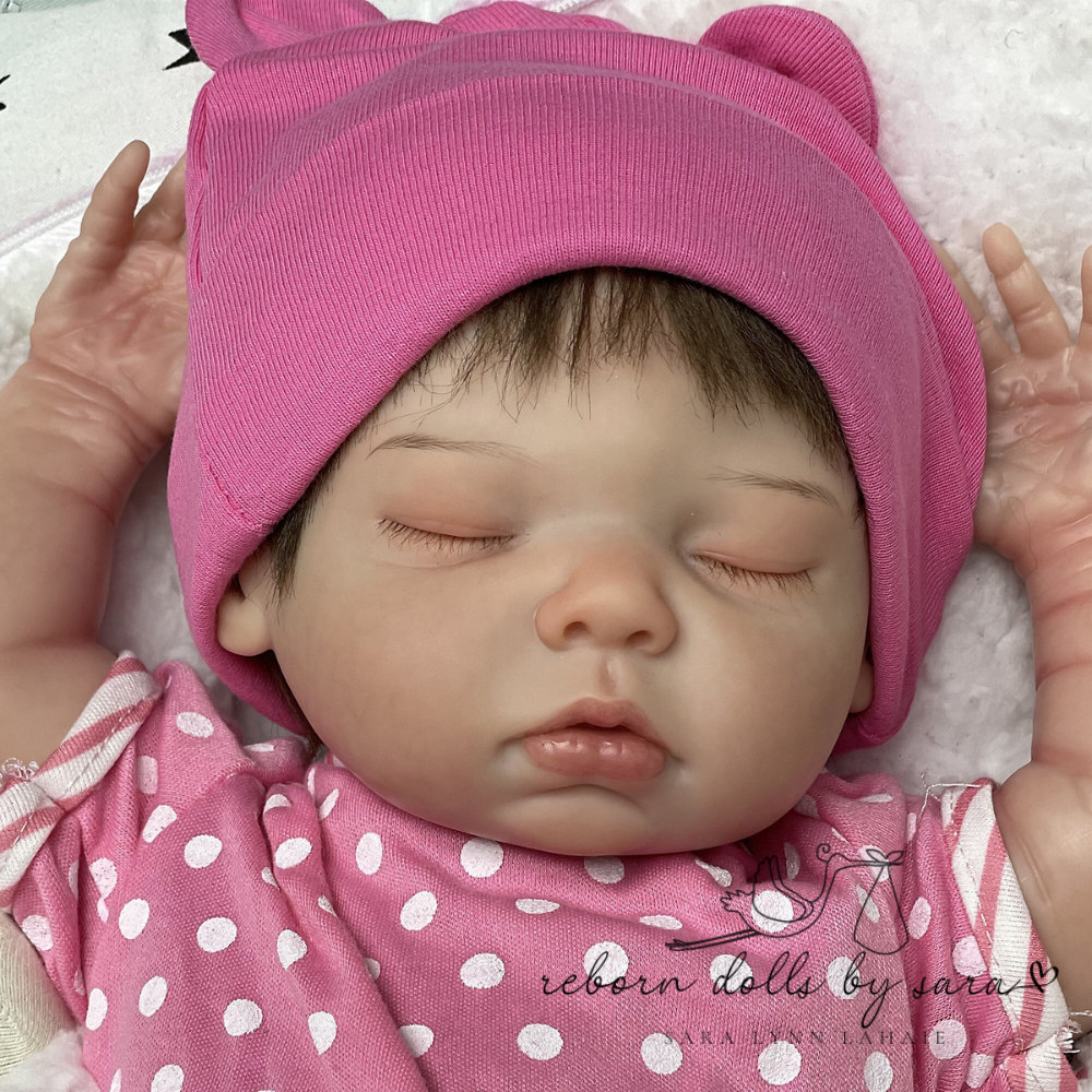 Cheap reborn baby girl doll Harlow asleep for sale by Reborn Dolls by Sara. Harlow is a preemie baby, 22" in length, asleep with brown hair and brown eyelashes. She's caucasian and wearing a pink and white outfit.