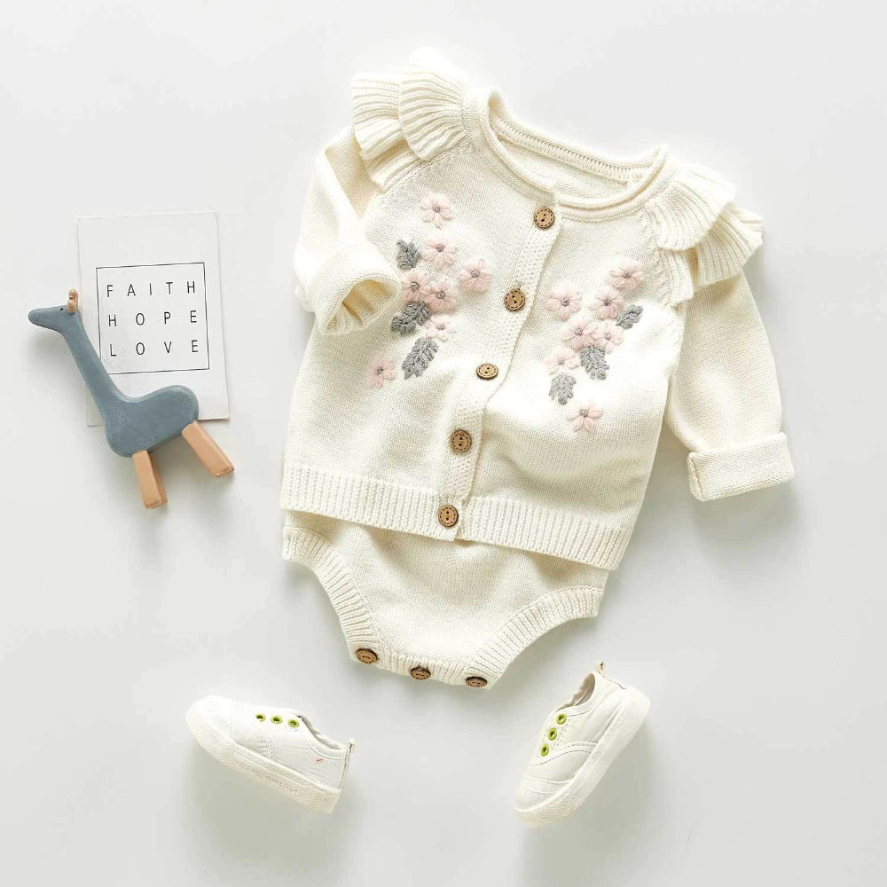 White knitted floral cardigan with frilly long-sleeves and buttons, and a matching overall onesie with button on straps and buttons at the crotch with pink and grey floral embroidery. Made for baby girls from newborn to 3 years old and reborn dolls.