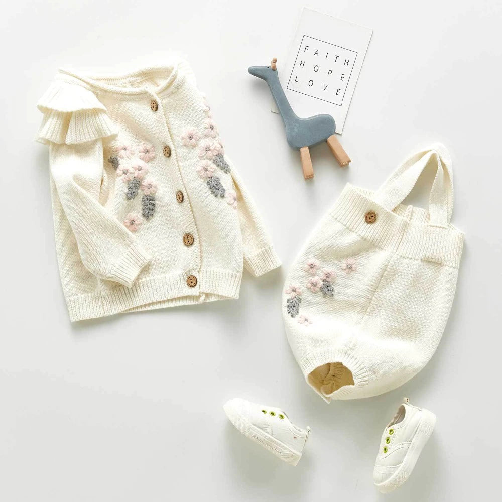 White knitted floral cardigan with frilly long-sleeves and buttons, and a matching overall onesie with button on straps and buttons at the crotch with pink and grey floral embroidery. Made for baby girls from newborn to 3 years old and reborn dolls.