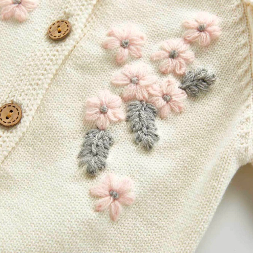 Close-up of the embroidery on the Creamy white knitted cardigan with frilly sleeves, long-sleeves, and buttons down the centre. Has beautiful pink and grey floral embroidery on the chest. Made for newborn baby girls to 3 years old and reborn dolls.