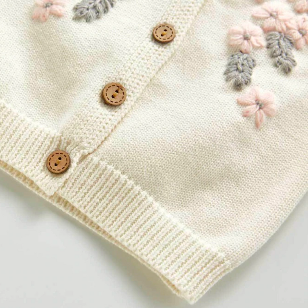 Close-up of the bottom hemline of the Creamy white knitted cardigan with frilly sleeves, long-sleeves, and buttons down the centre. Has beautiful pink and grey floral embroidery on the chest. Made for newborn baby girls to 3 years old and reborn dolls.